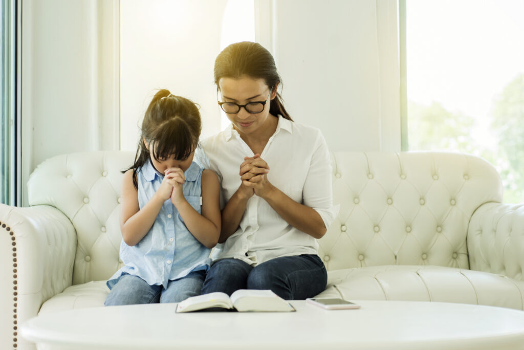 A mother and young daughter sitting on a couch and praying over a Bible while waiting patiently on God and his promises.