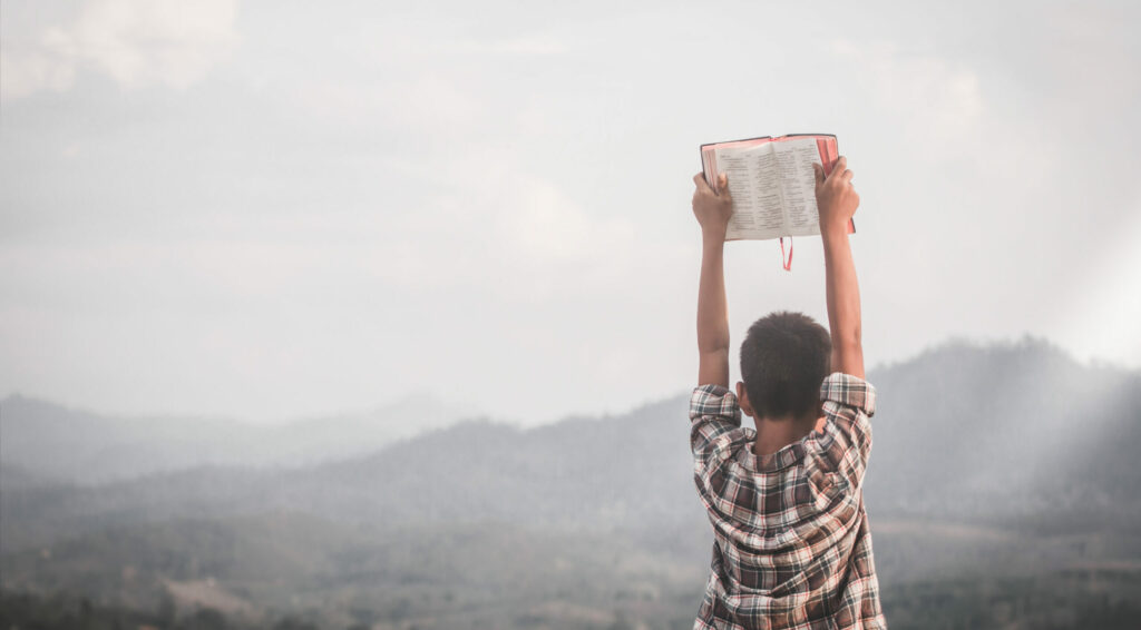 A young boy raises the bible up in front of a landscape.