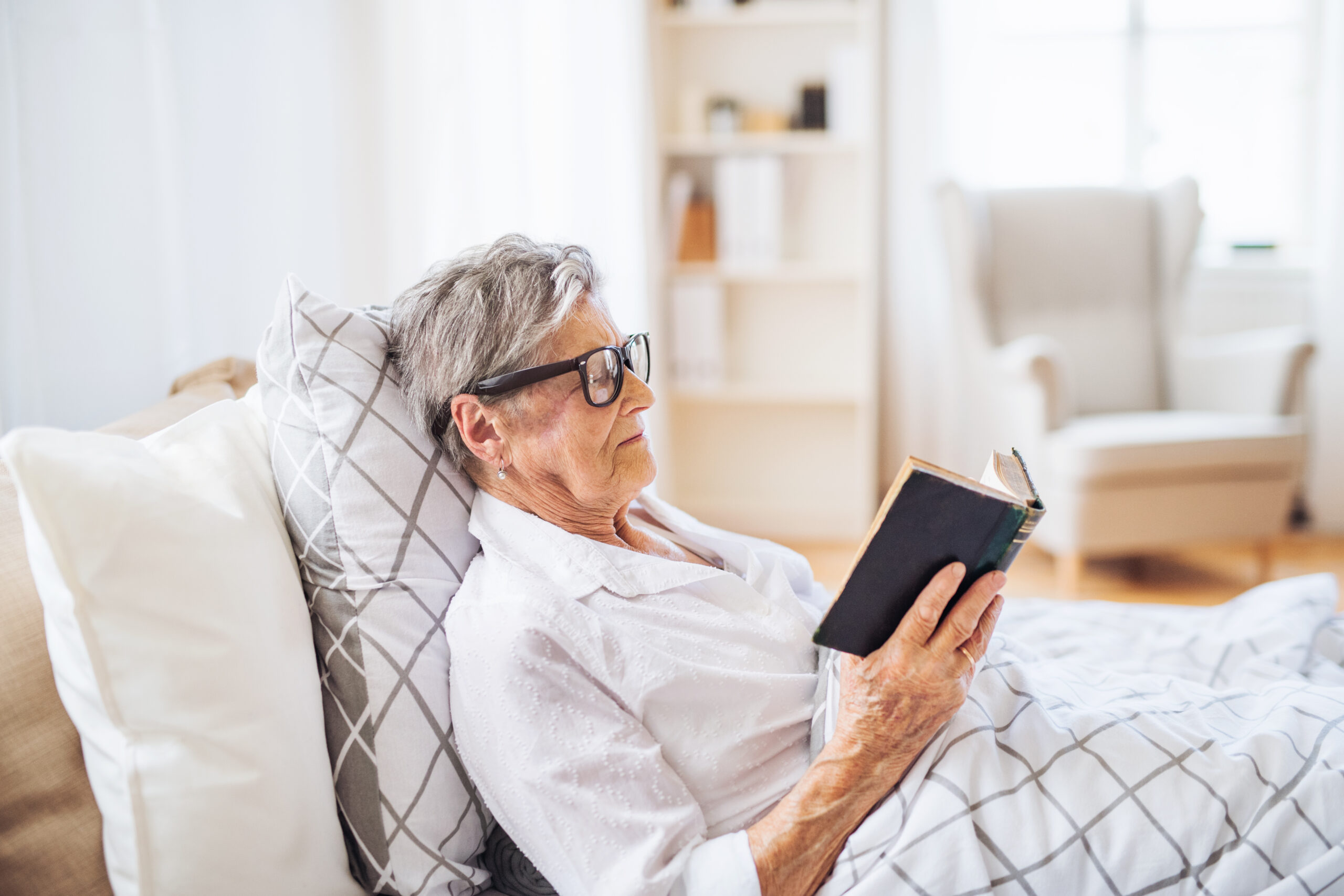 A senior woman with eyeglasses lying in bed at home reading the bible.