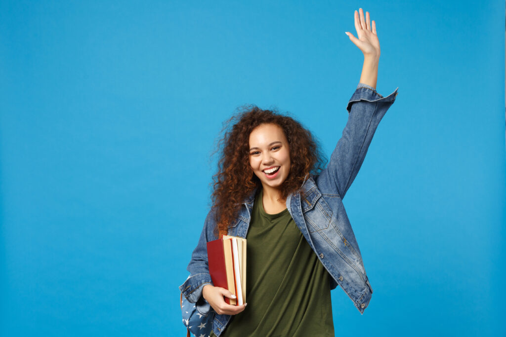 Teenage girl wearing jean jacket and carrying books with one arm raised in joy in front of blue background.