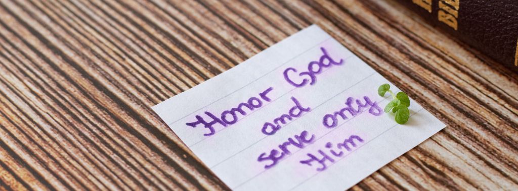 A handwritten note in purple resting on a desk with the message honor God and serve only Him next to a Bible.