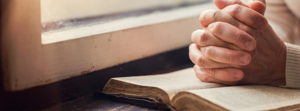 Woman with hands together, praying over Bible next to window.