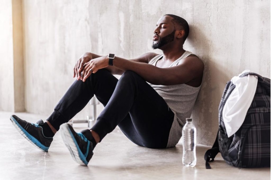 Man wearing athletic clothing resting against concrete wall, sitting next to plastic water bottle and backpack.