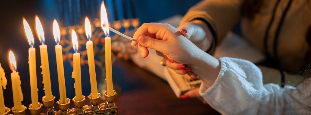 Woman's hand guiding child's hand to light candles on Menorah with glittery background.