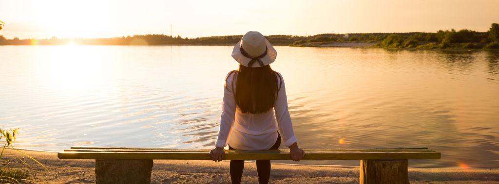  A young woman in a hat sitting on a bench facing a lake at sunset. 
