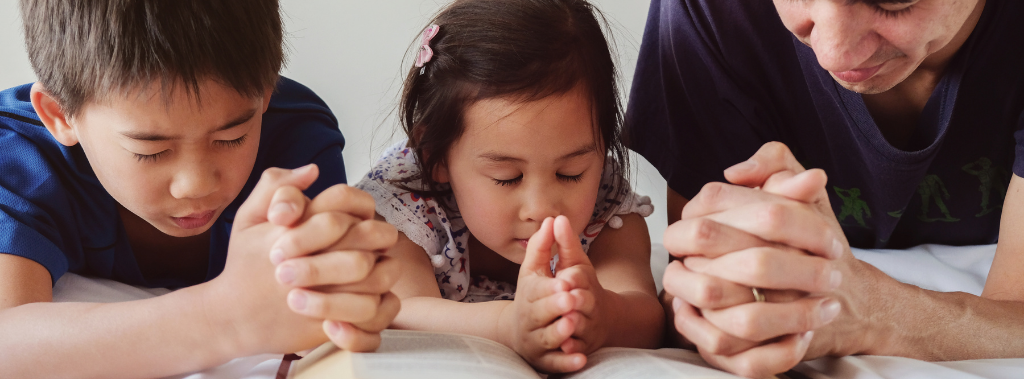 A father, son, and daughter praying and reading from a Bible
