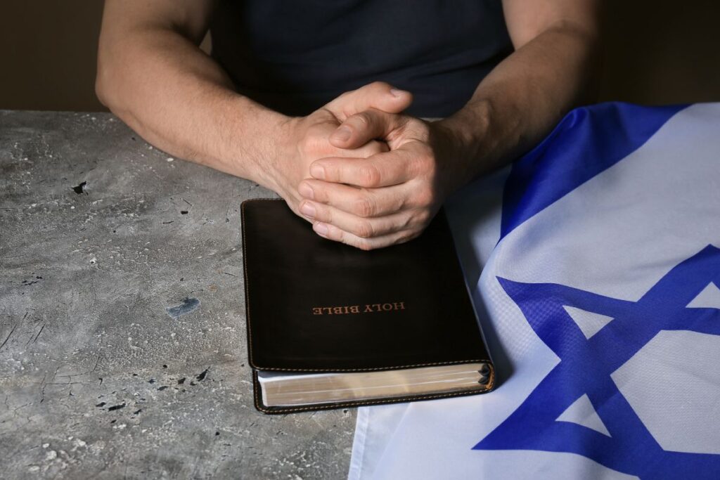 Hands folded in prayer resting on top of the Holy Bible with the flag of Israel laying under the Bible.